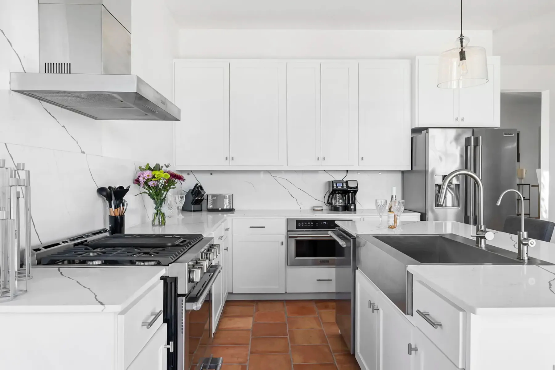 Modern kitchen with white cabinets, marble countertops, stainless steel appliances, and a vase of flowers. A large sink and tile flooring are visible.