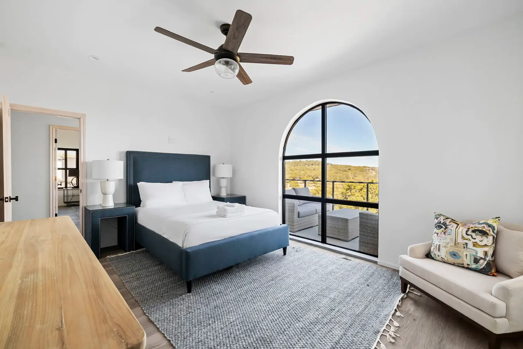 Modern bedroom with a blue upholstered bed, large arched window, ceiling fan, wooden dresser, and an armchair with a colorful pillow.
