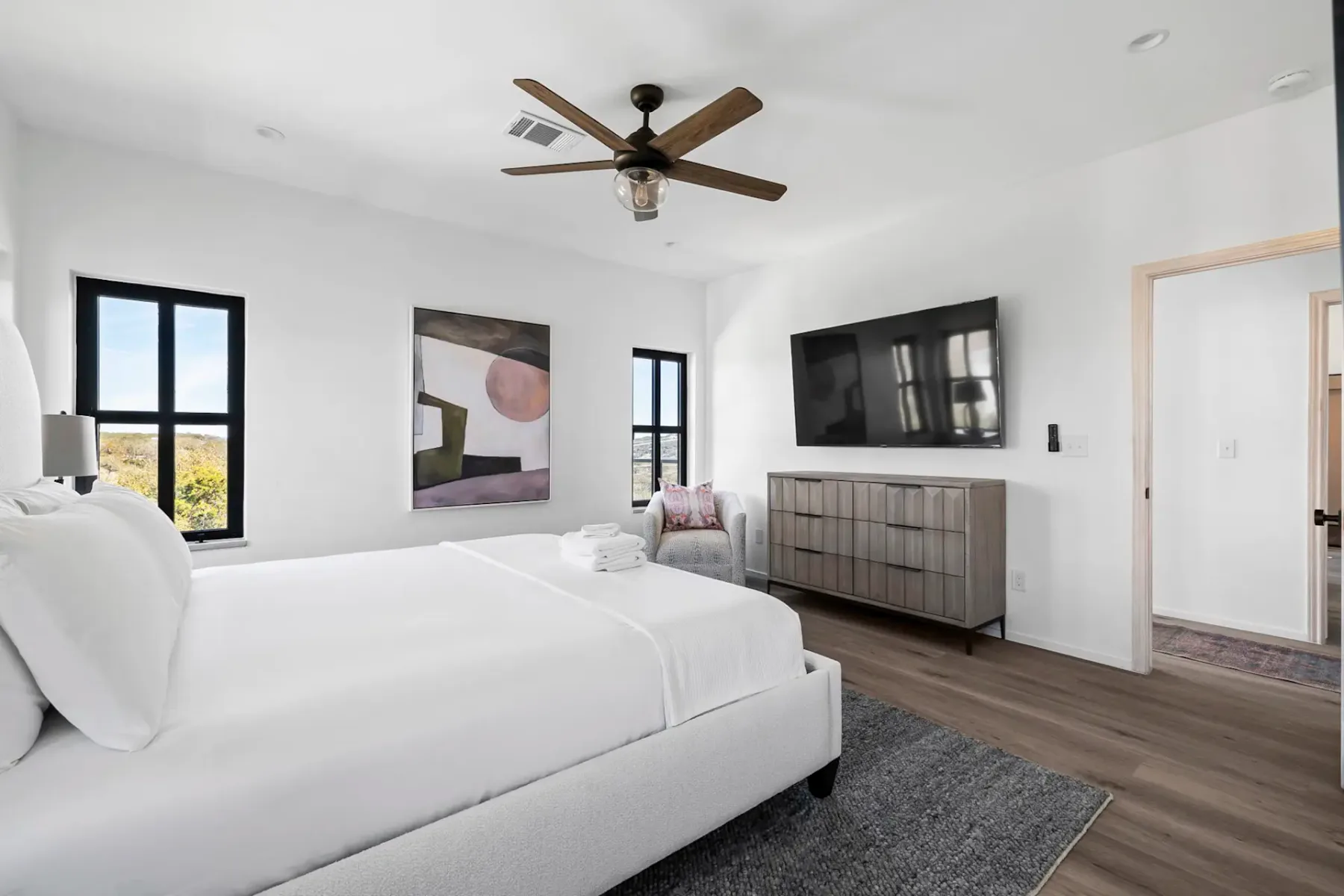 Minimalist bedroom with a white bed, wooden dresser, wall art, ceiling fan, and large TV. Bright windows and hardwood floor enhance the space.