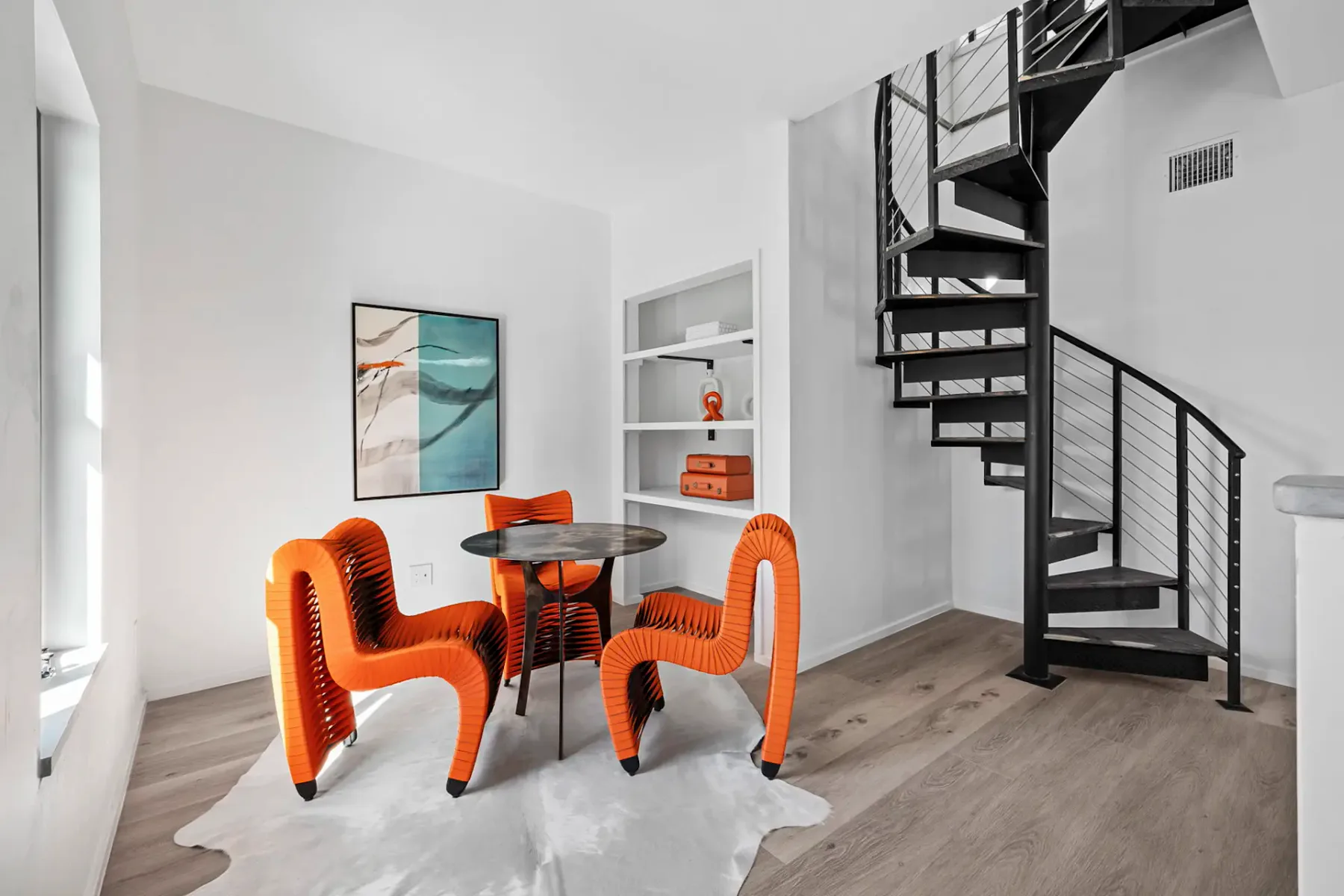 Modern dining area with three orange chairs around a round table, abstract artwork on the wall, and a spiral staircase in the background.