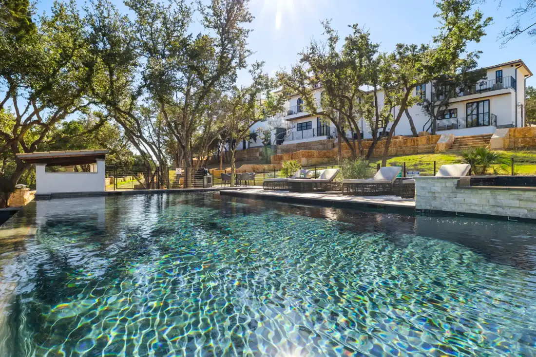 Luxurious swimming pool with clear water reflecting sunlight, surrounded by lounge chairs, trees, and a large white house in the background.