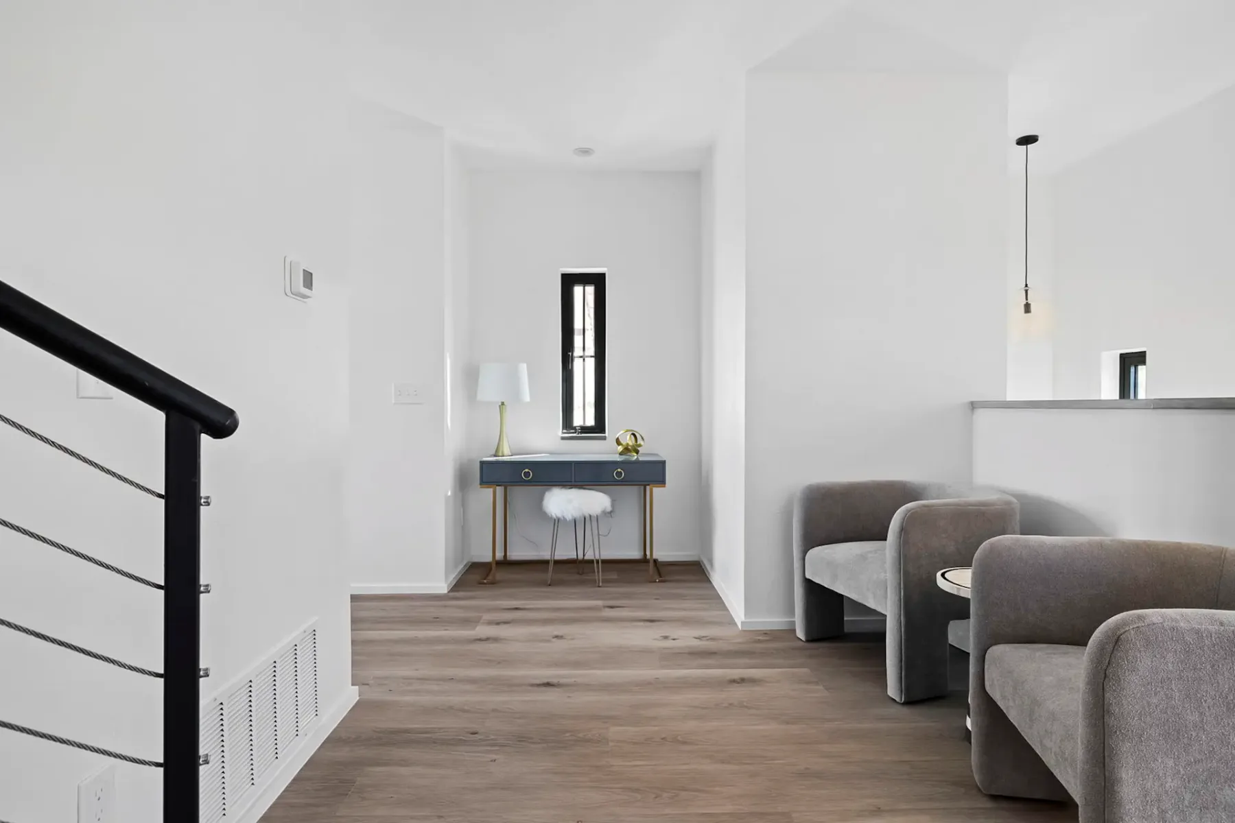 Habitación minimalista con dos sillones grises, un pequeño escritorio con una lámpara y una flor, suelo de madera y una lámpara colgante. Las paredes blancas aportan luminosidad.
