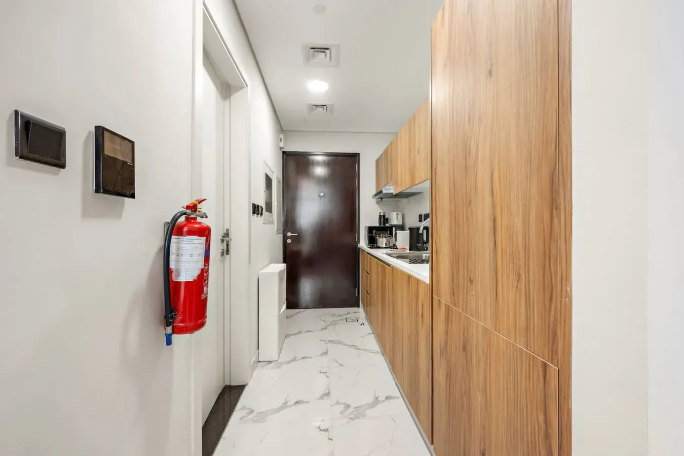 Un pasillo estrecho con armarios de madera y una pequeña cocina conduce a una puerta oscura.