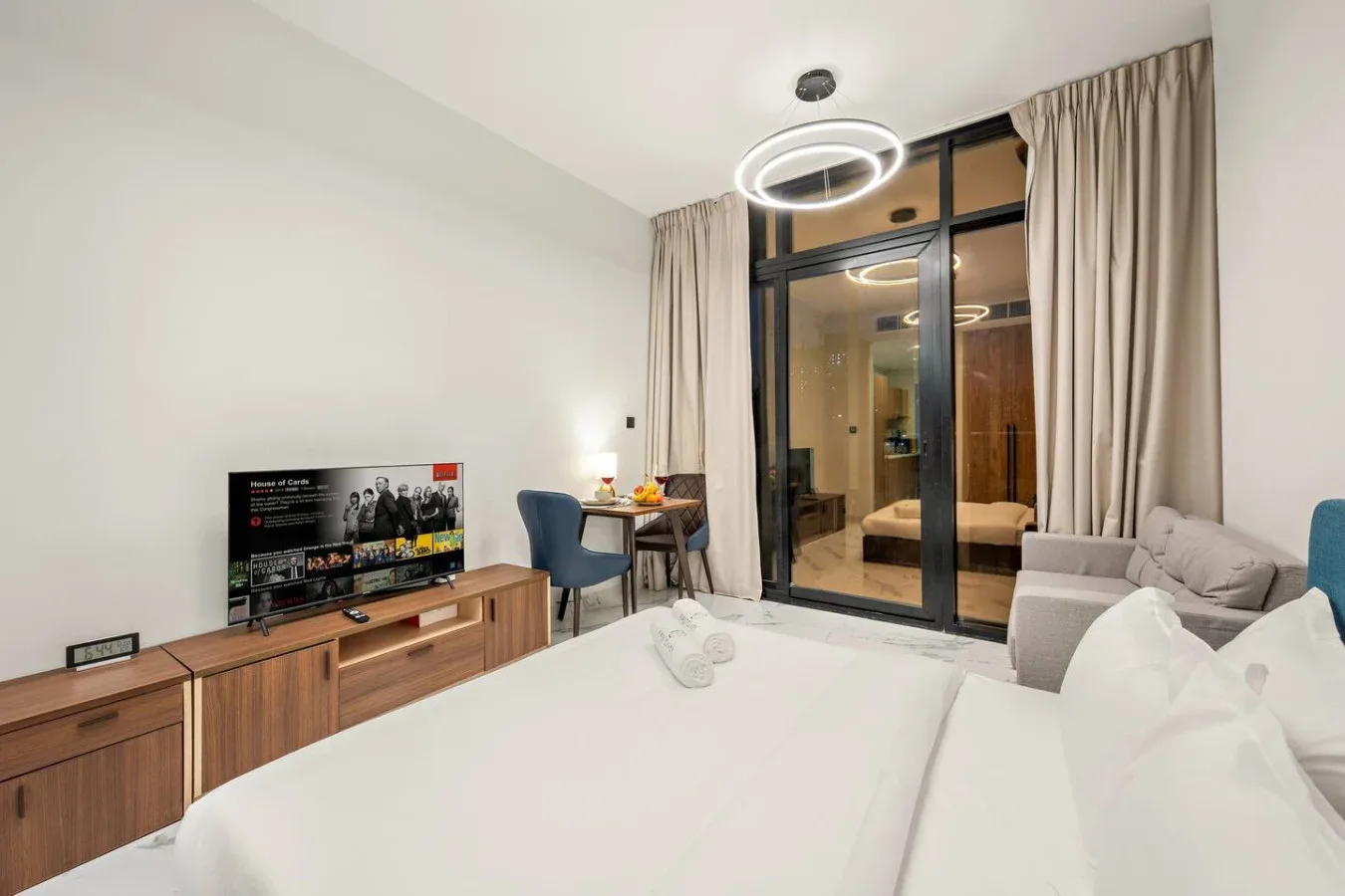 Un dormitorio con una cama blanca, televisor y puertas de vidrio junto a cortinas beige.