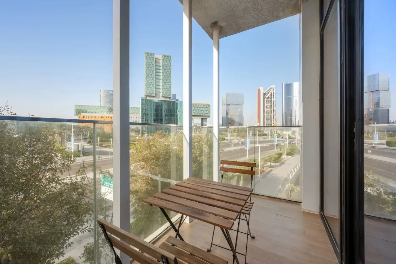 A balcony with a wooden table and seating overlooks nearby modern buildings.