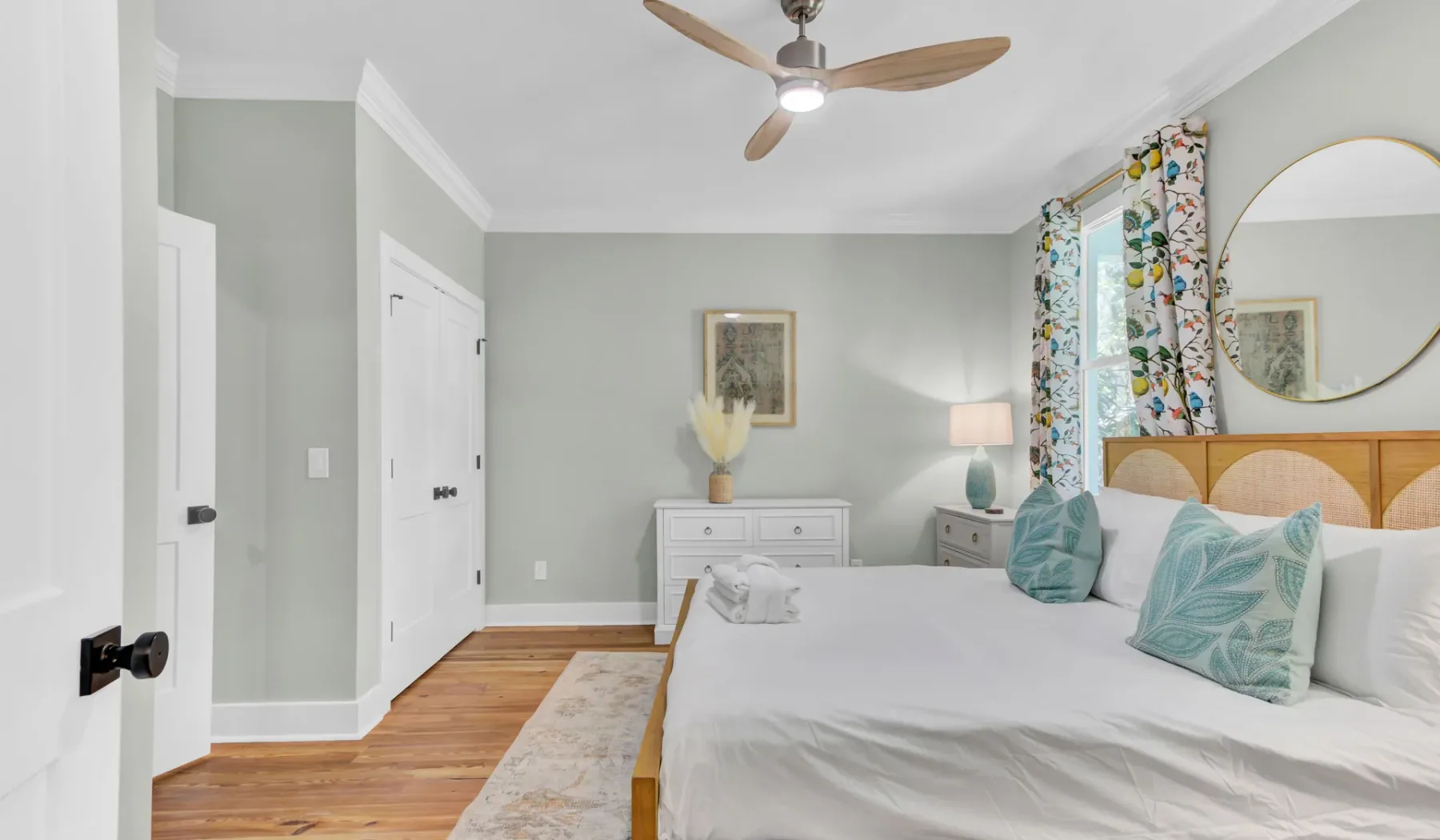 A bright bedroom with a white bed, teal pillows, and a ceiling fan.