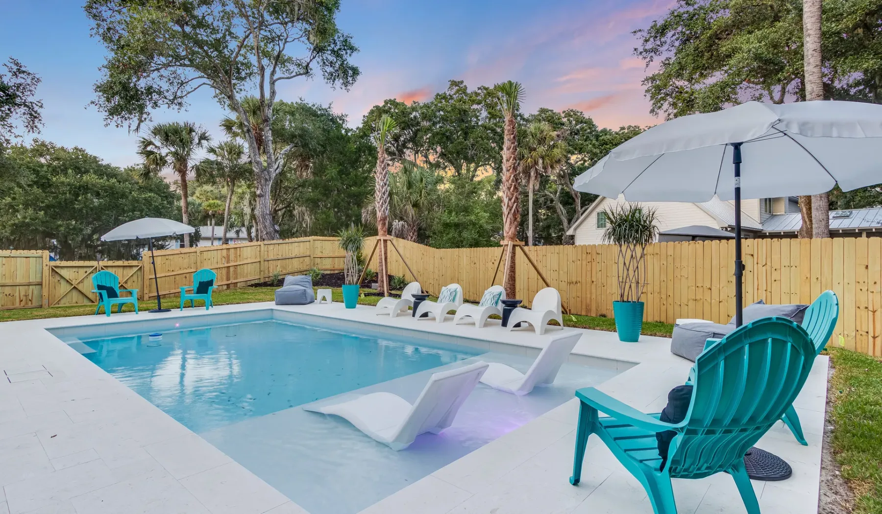 An outdoor swimming pool is surrounded by lounge seating, umbrellas, and a wooden fence.