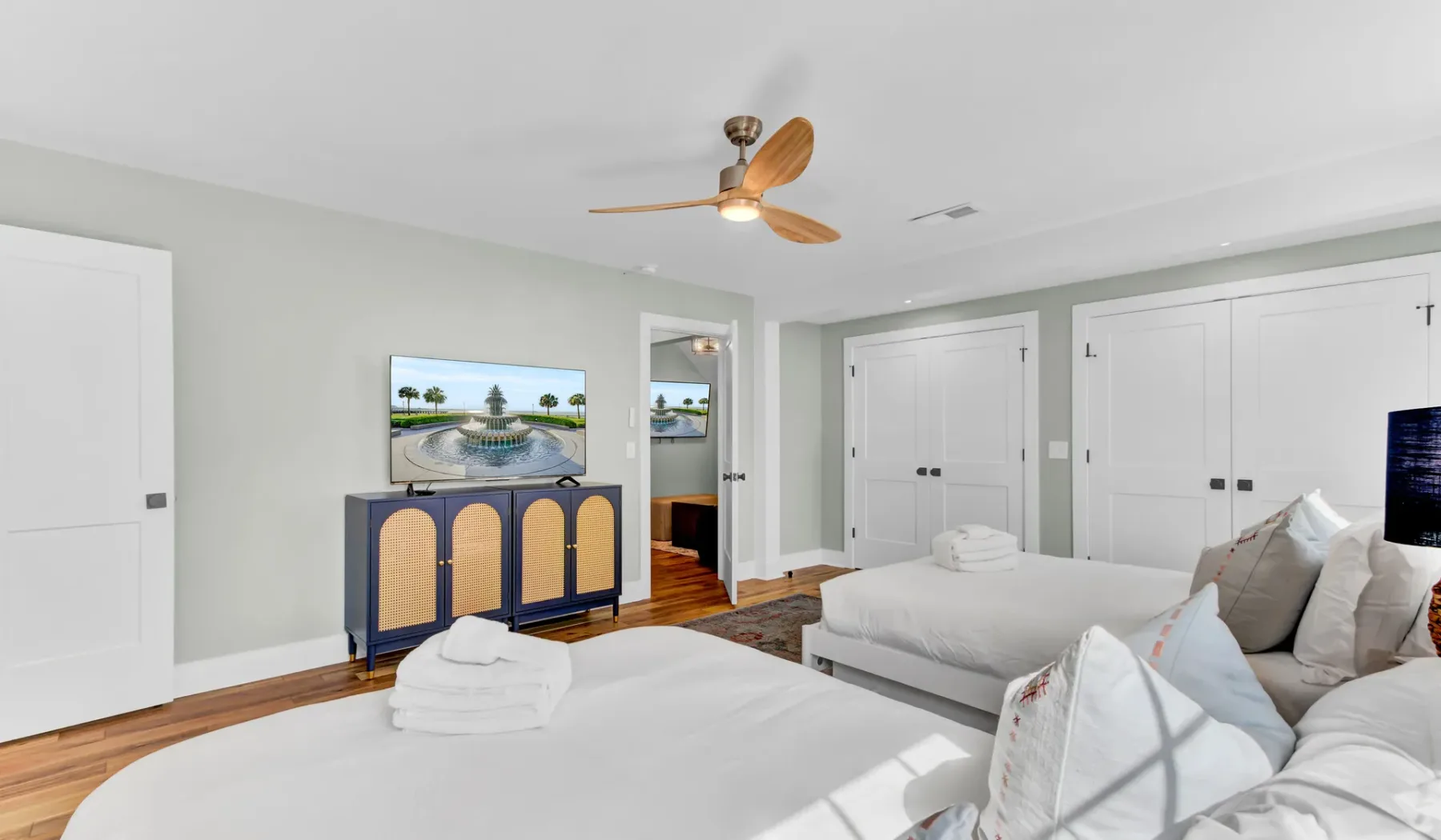 A bright bedroom with white bedding, a ceiling fan, and a television on a cabinet.