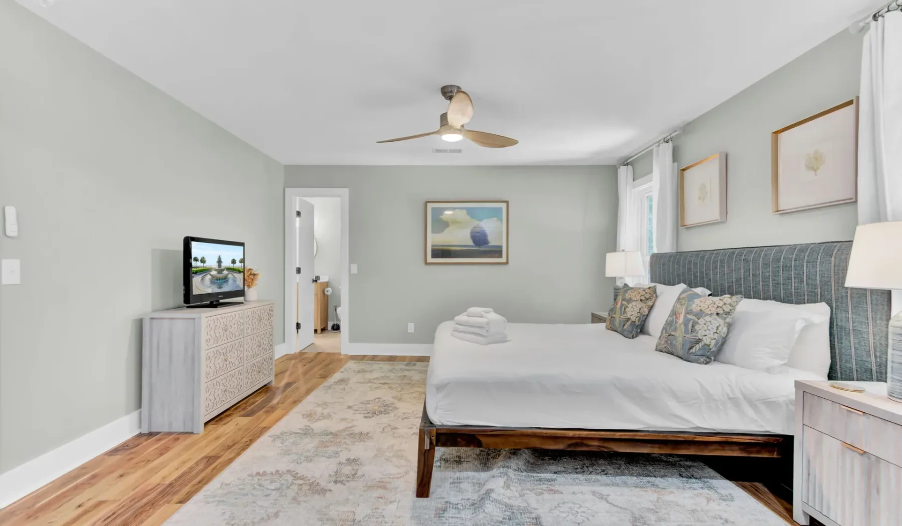 A bright bedroom with a large bed, framed artwork, and a television on a dresser.