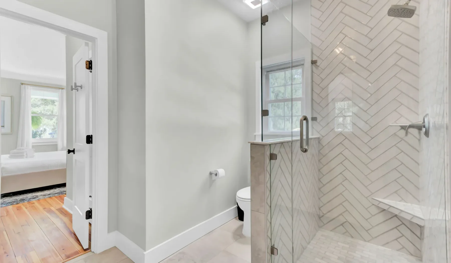 A bright bathroom with a glass shower, toilet, and doorway to a bedroom.