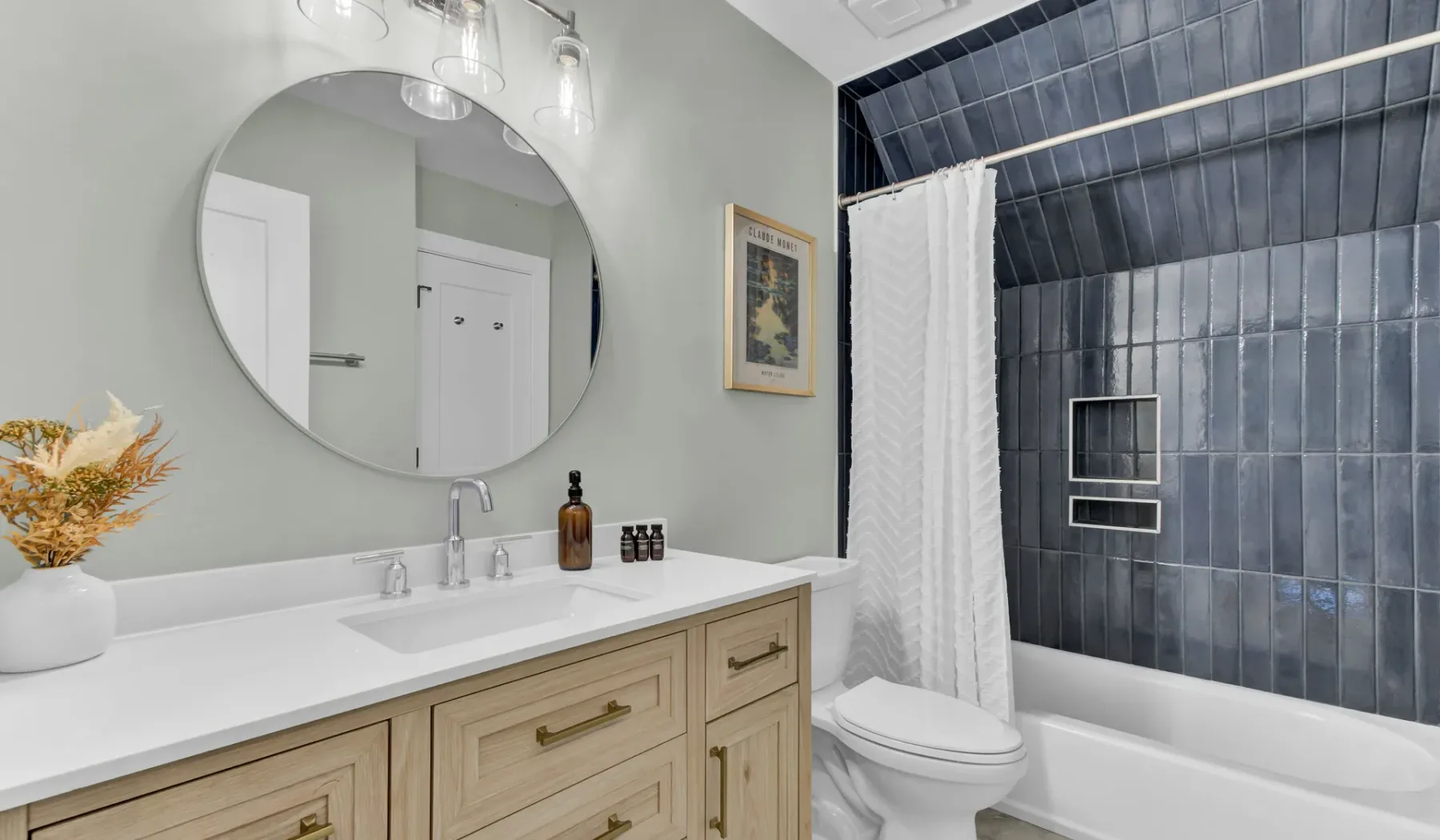 A bright bathroom with a long vanity, round mirror, and bathtub.