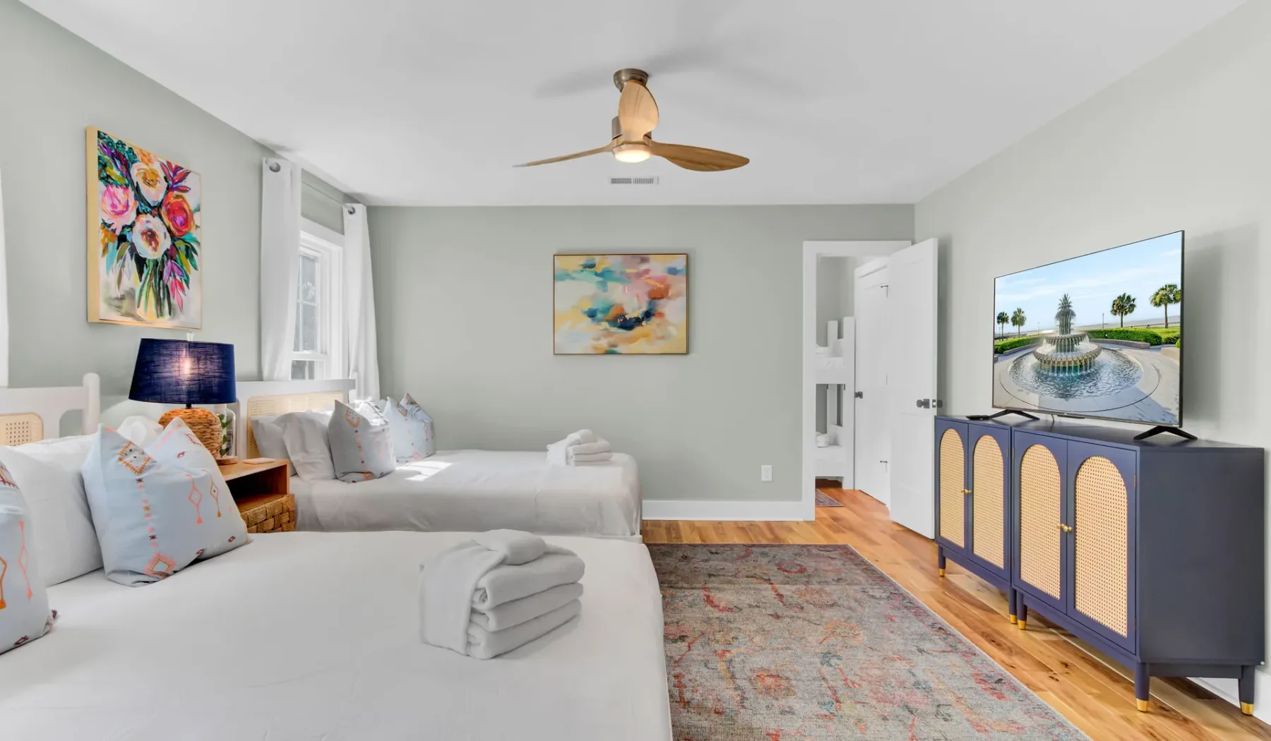 A bright bedroom with two beds, colorful artwork, and a television.