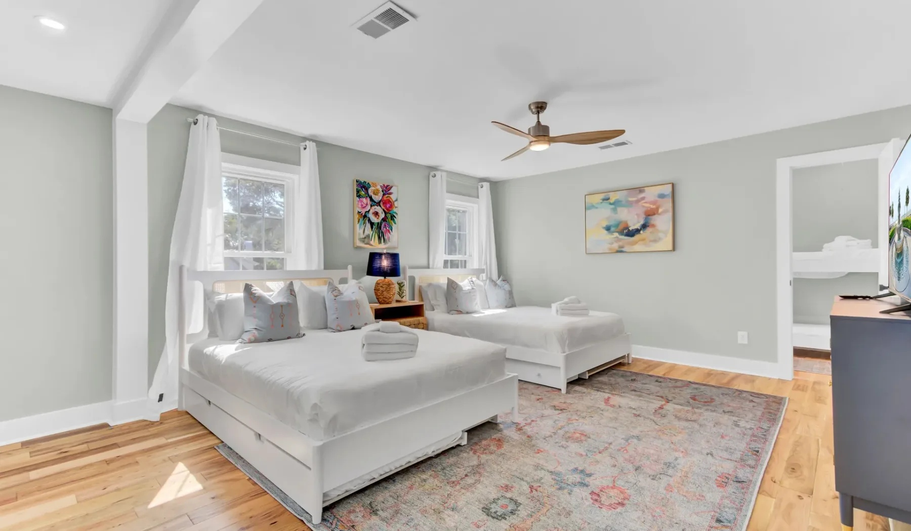 A bright bedroom with two beds, a patterned rug, and windows with curtains.