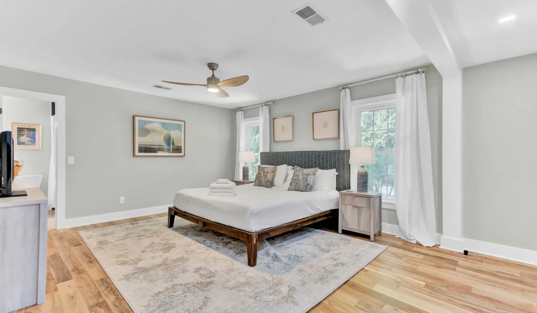 A bright bedroom with a neatly made bed, bedside table, and large windows.
