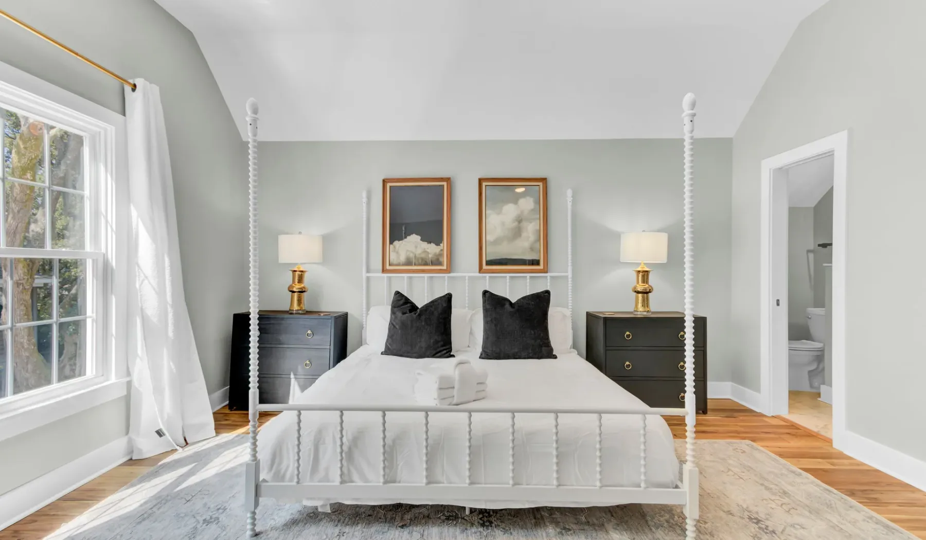 A bright bedroom with a white bed, dark pillows, and framed artwork above the headboard.