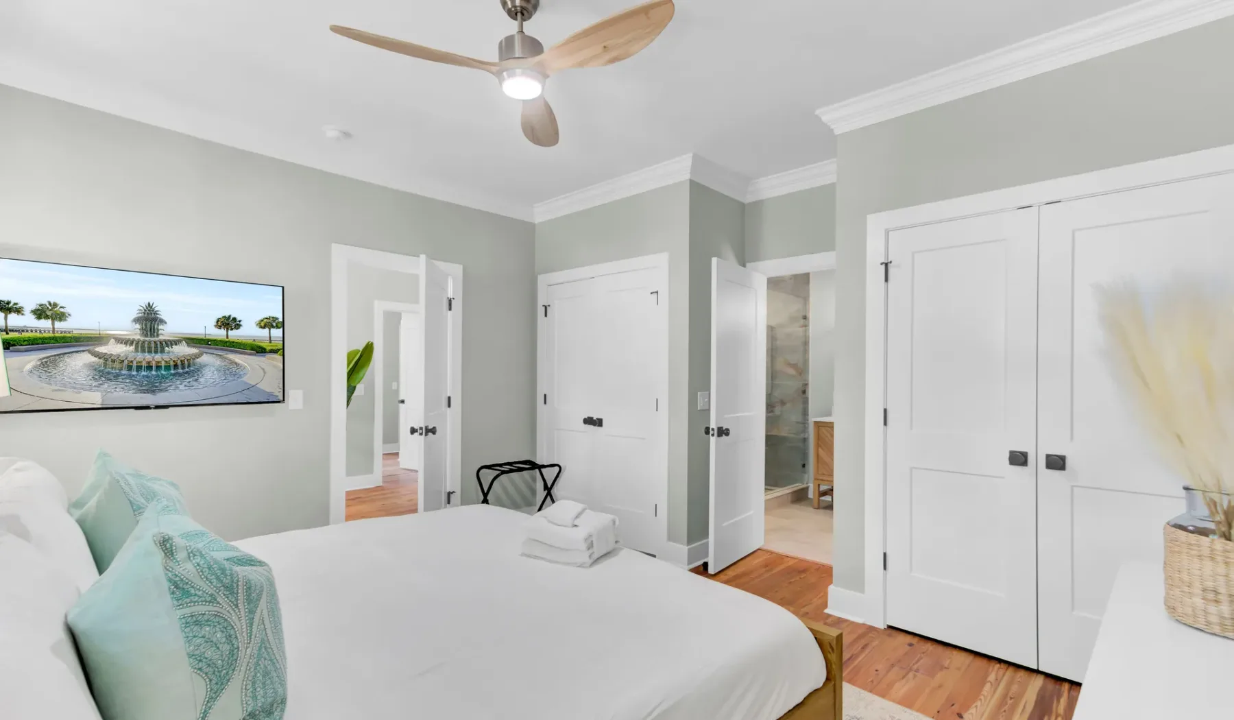 A bright bedroom with a white bed, pale green walls, and hardwood flooring.