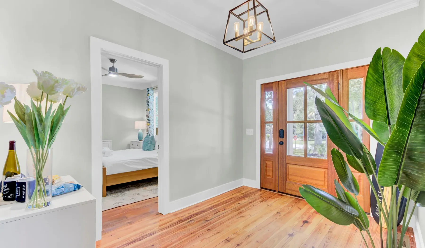 A bright entryway with a wooden front door, a potted plant, and a bedroom visible through the doorway.