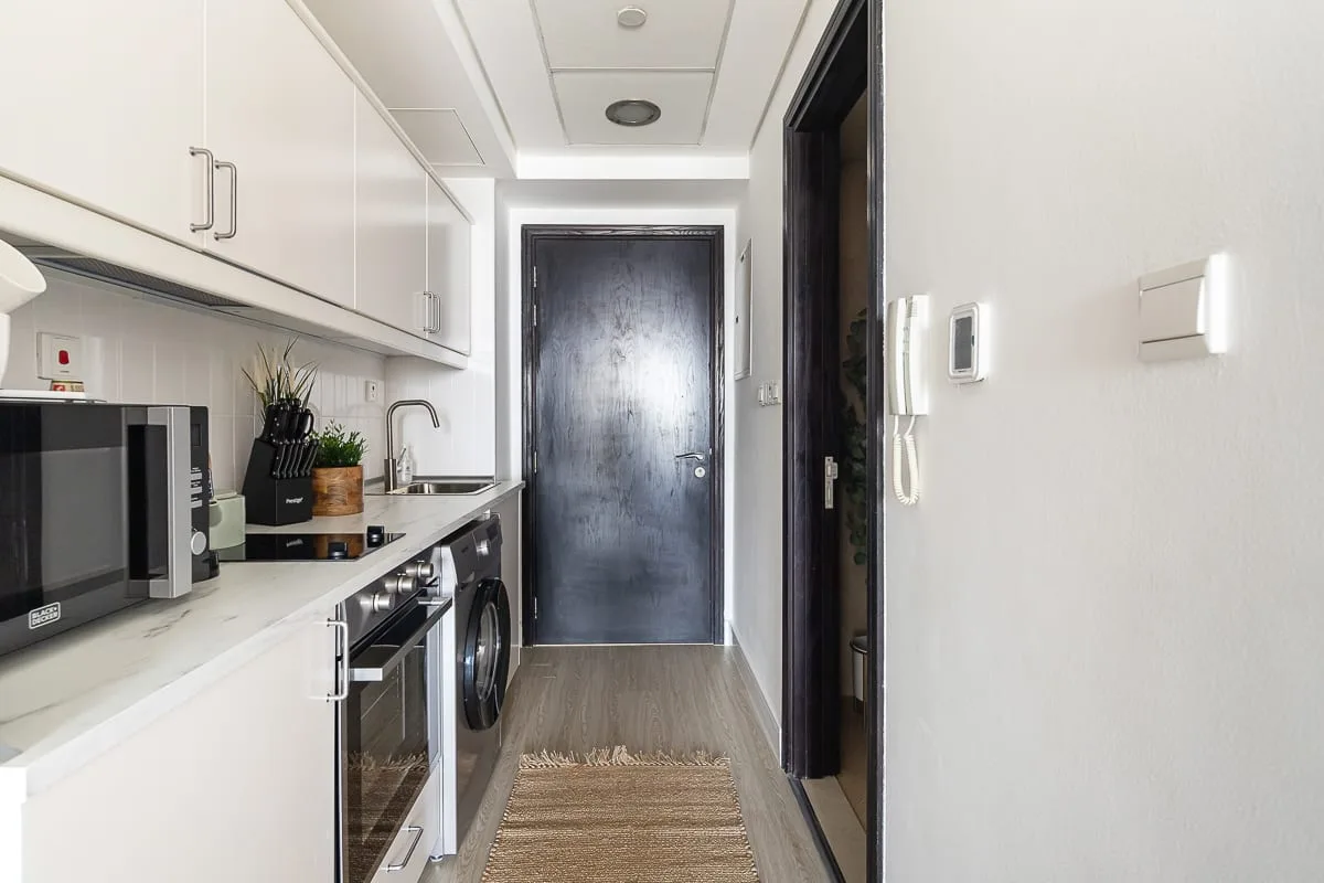Una cocina compacta con gabinetes blancos y una puerta oscura al final del pasillo.