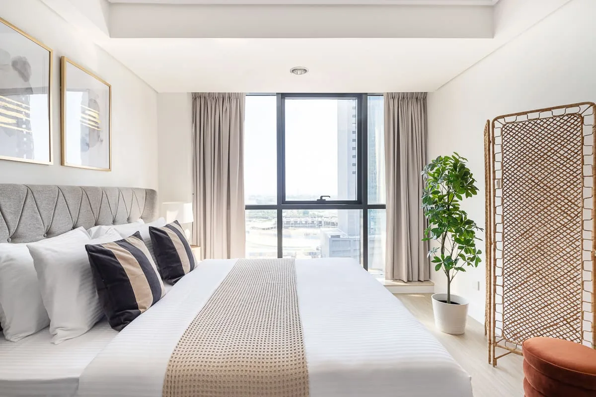 Un dormitorio luminoso con una cama bien tendida, una ventana grande y una planta en maceta.