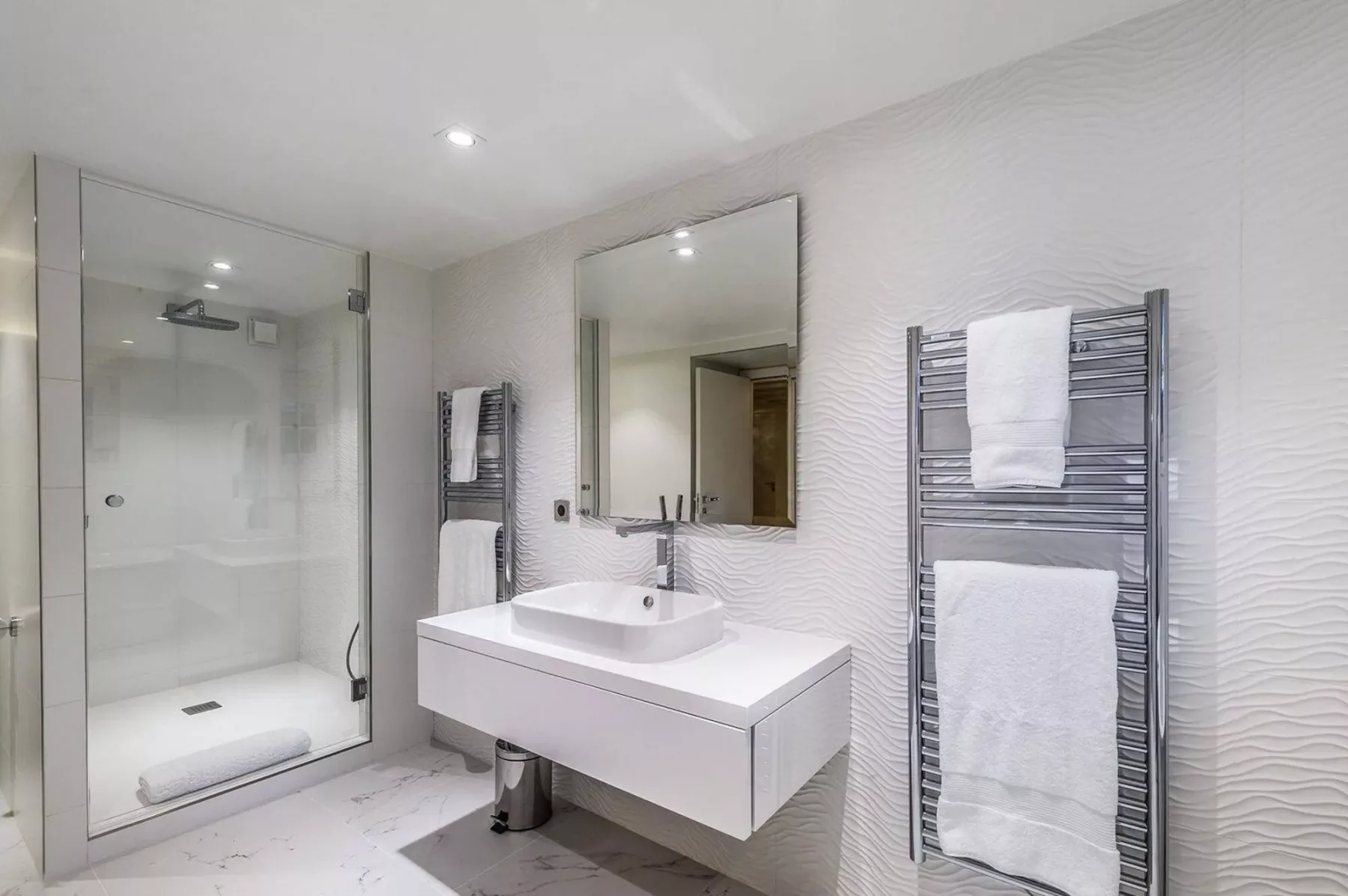 Un baño luminoso con ducha de vidrio, espejo de pared y lavabo suspendido.
