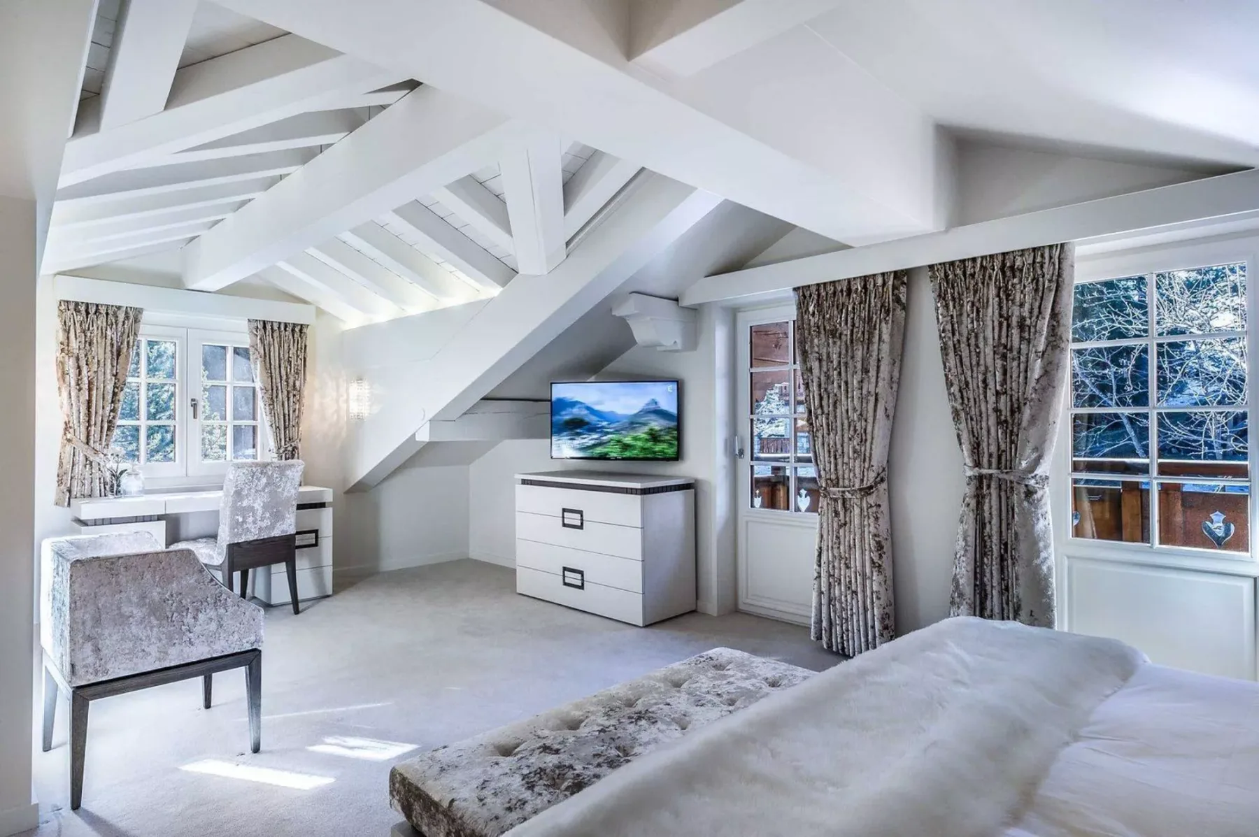 Un dormitorio luminoso con cama, cómoda y grandes ventanas bajo vigas blancas expuestas.