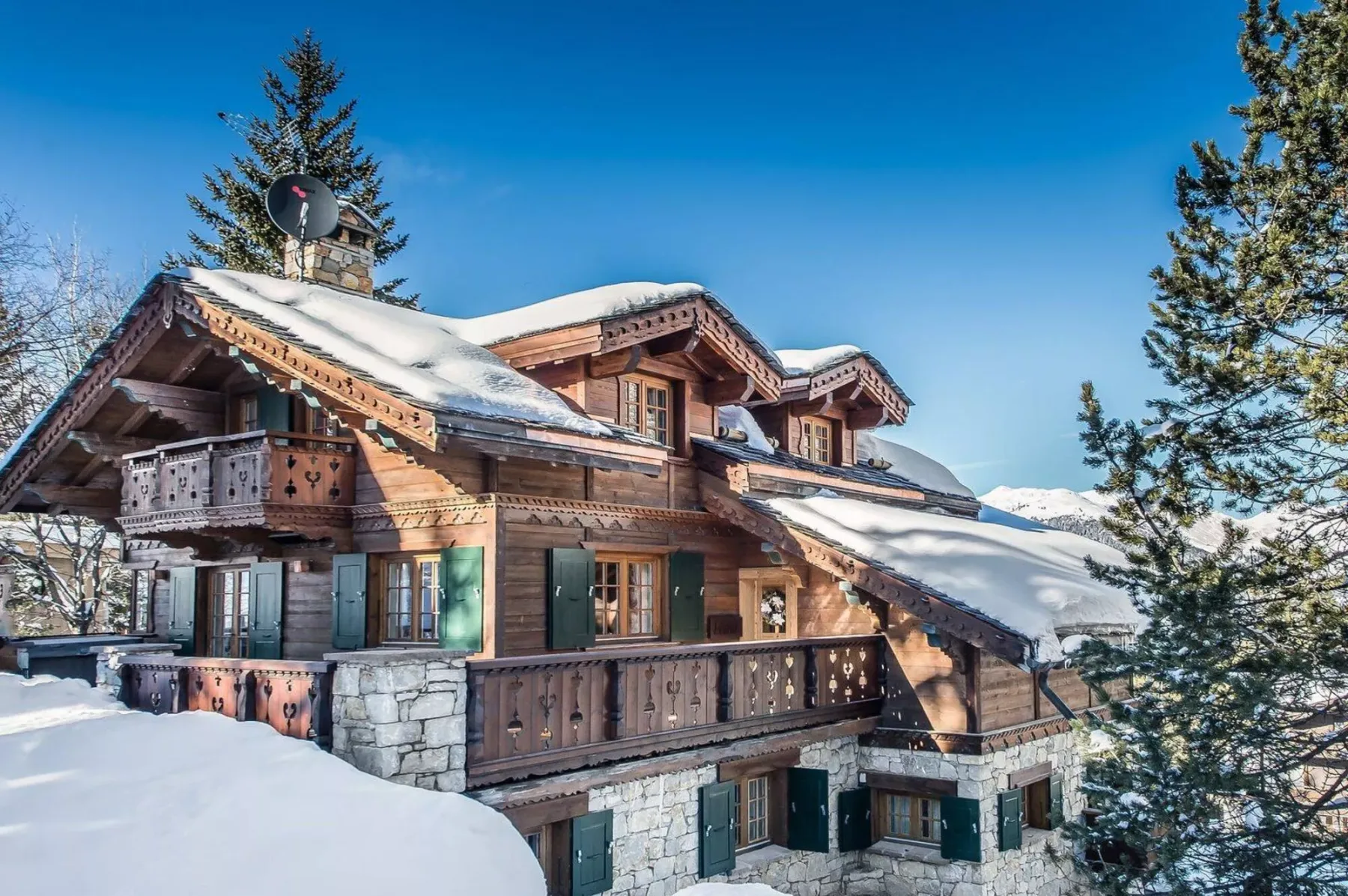 Un chalet cubierto de nieve, con balcones de madera, está entre árboles perennes.