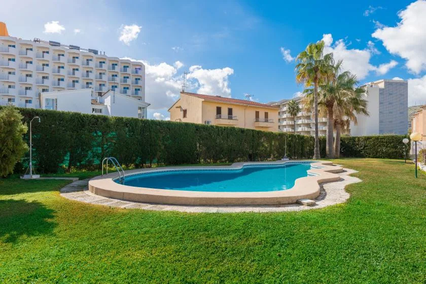 An outdoor swimming pool sits in a grassy courtyard beneath a bright blue sky.