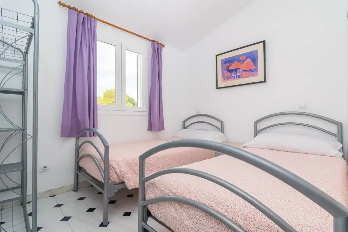 A bedroom with metal-framed beds, pale bedding, and a window with purple curtains.