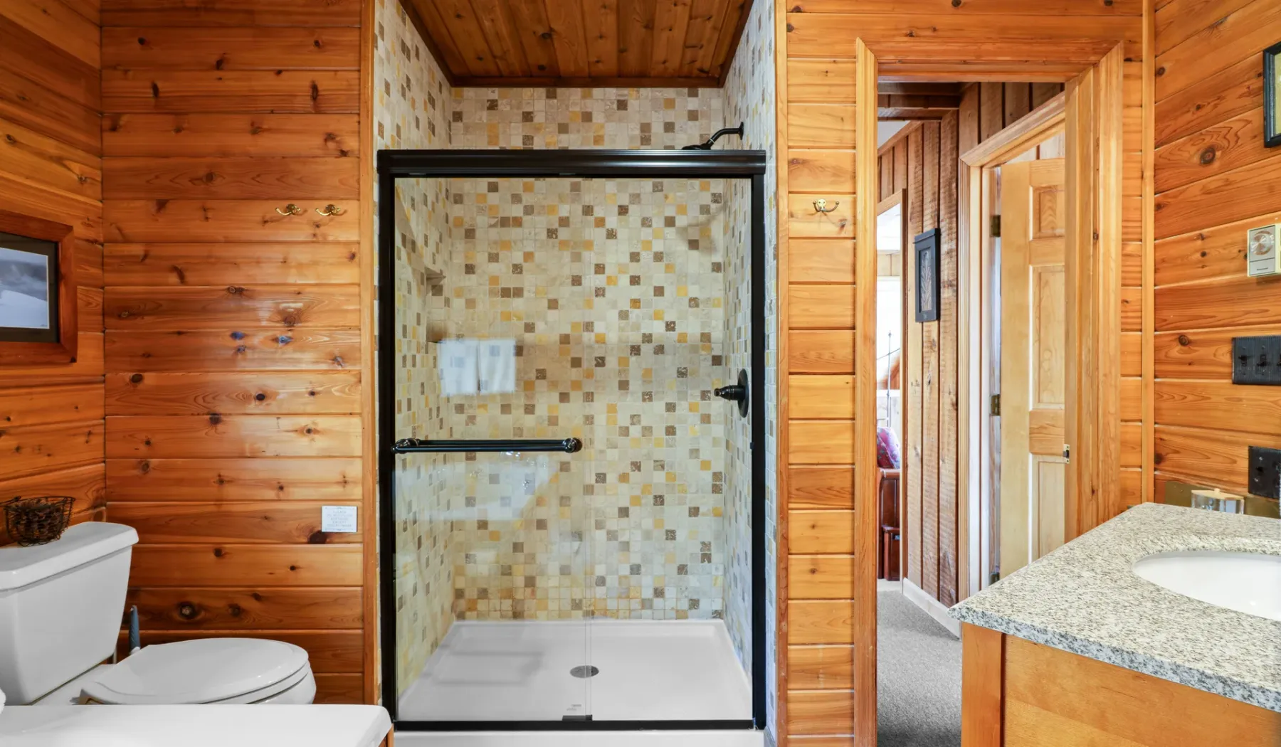Un baño con ducha de vidrio, inodoro y tocador con lavabo, rodeado de paredes revestidas de madera.