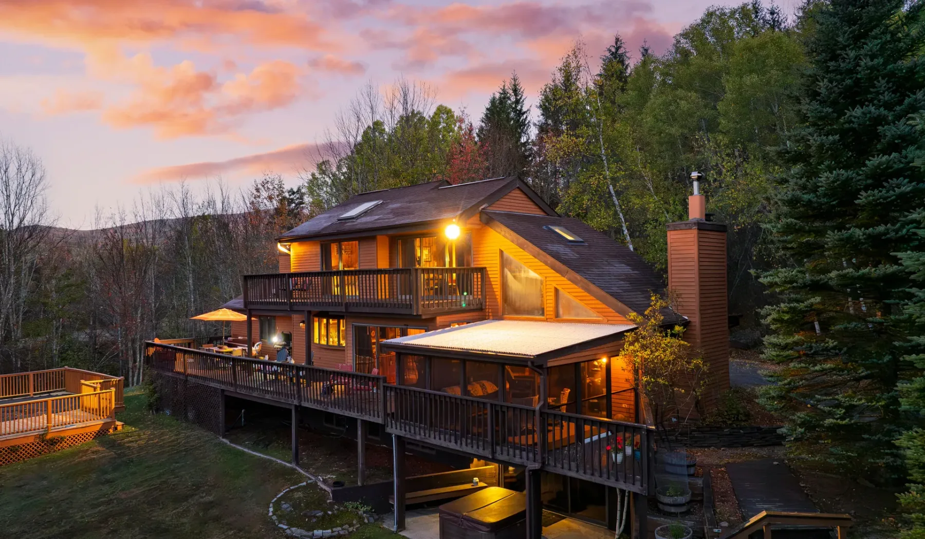 Una casa de madera iluminada, con amplias terrazas, se encuentra entre árboles al atardecer.