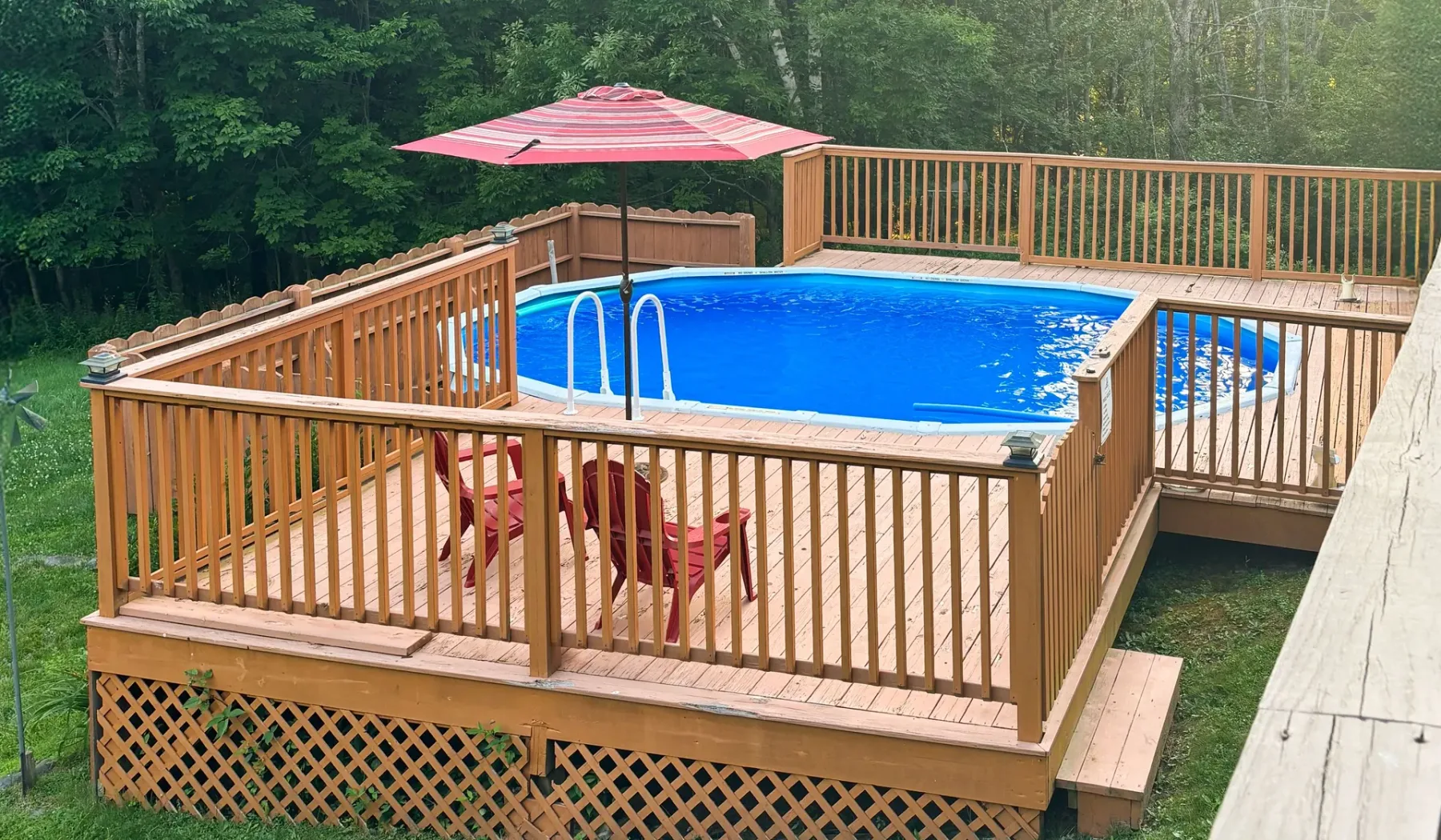 Una piscina elevada al aire libre está rodeada de barandillas de madera, con una sombrilla roja cerca.