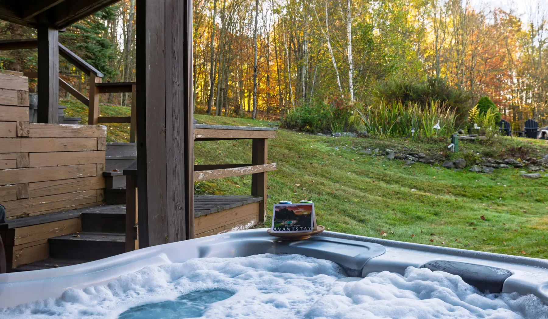 Una bañera de hidromasaje al aire libre está junto a una terraza de madera, con árboles otoñales al fondo.