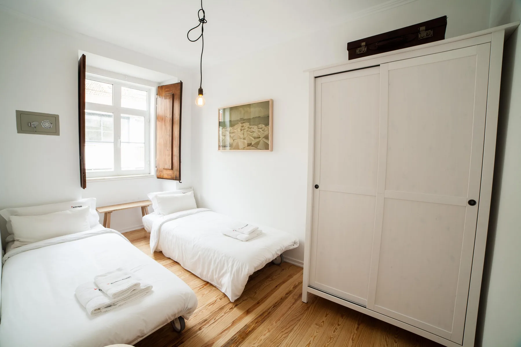 A bright bedroom with two beds, a window, and a large wardrobe.