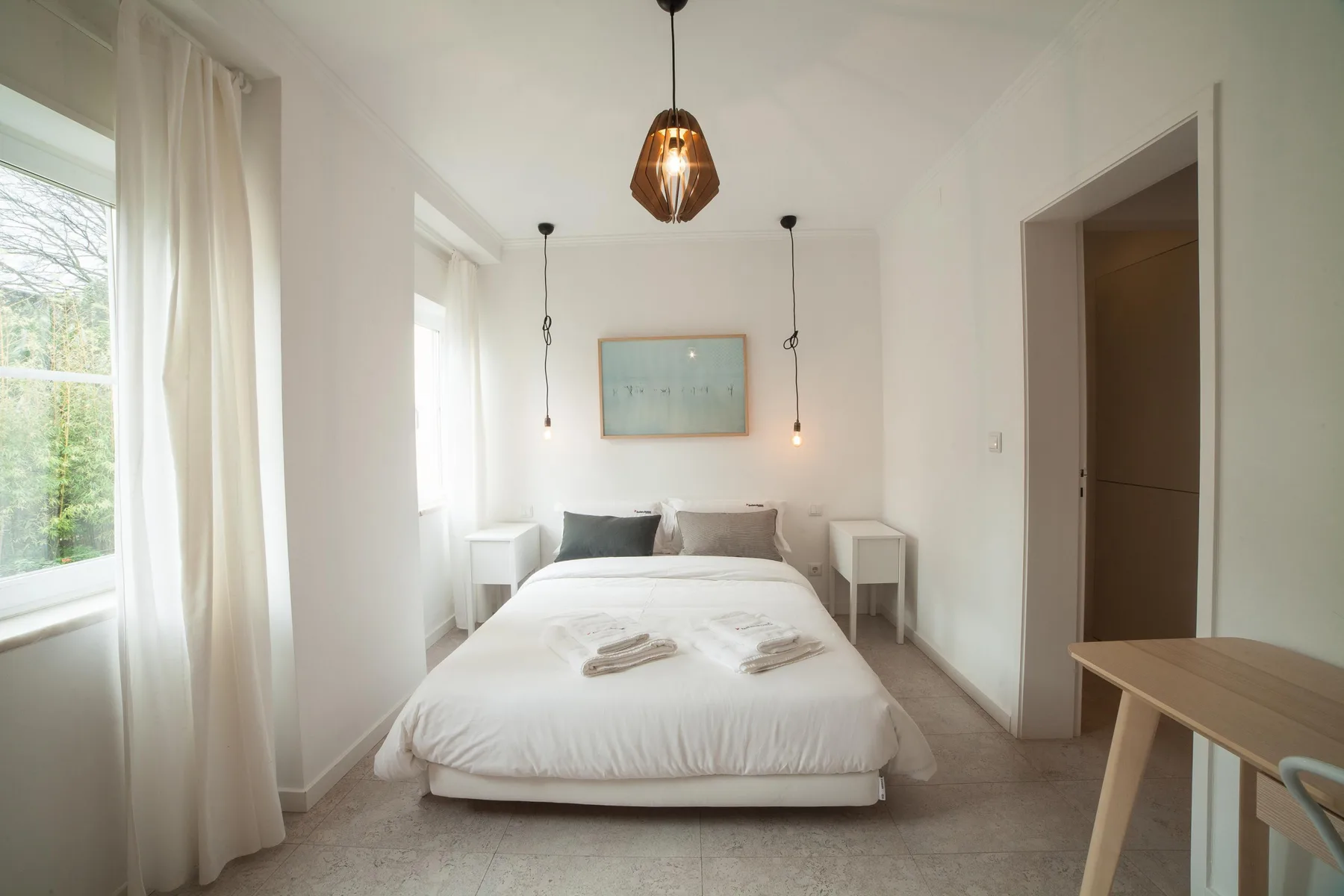 A bright bedroom with a bed, bedside tables, and large windows.