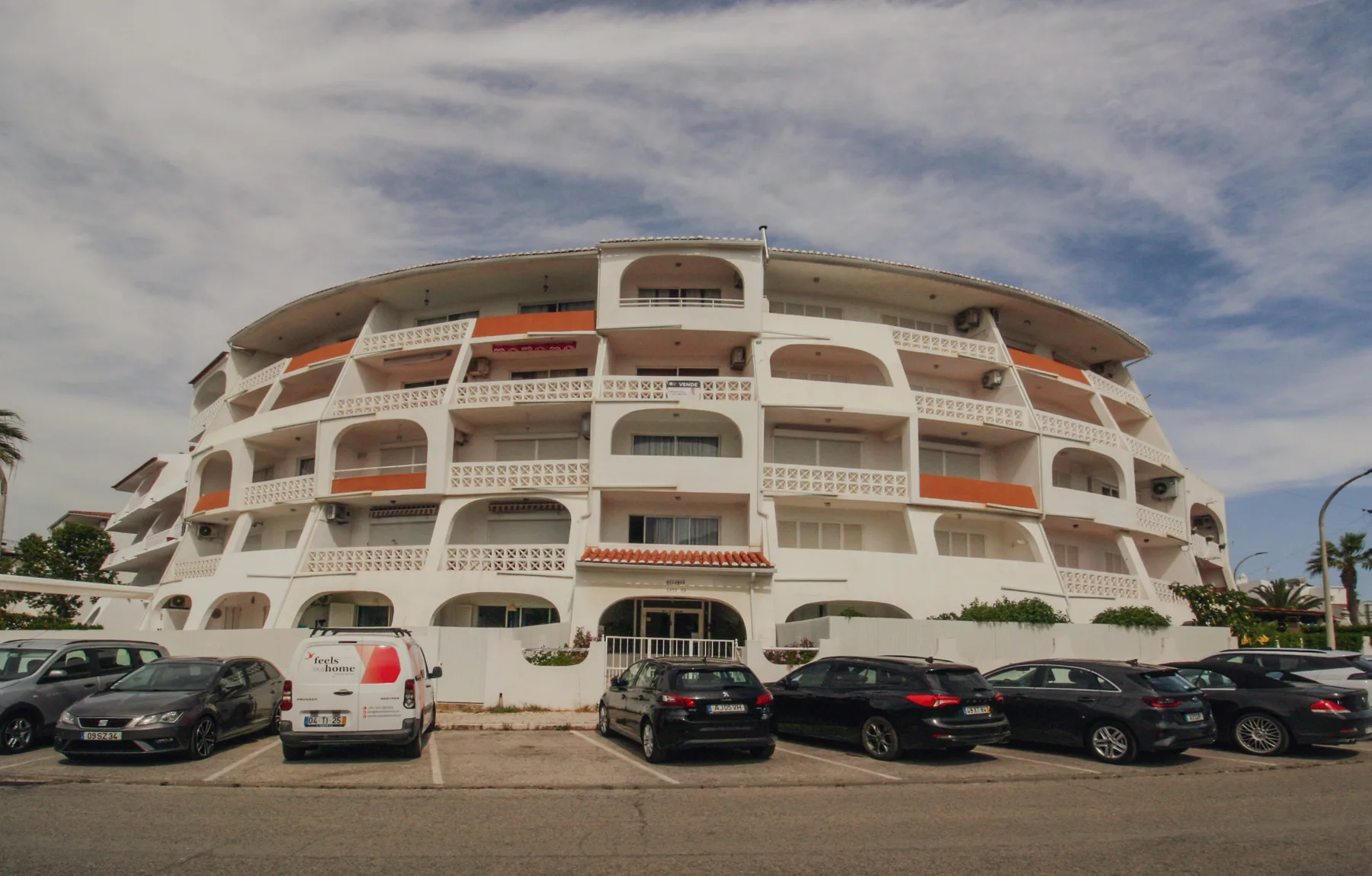 Un edificio de apartamentos curvo con balcones se alza junto a una calle con coches estacionados.