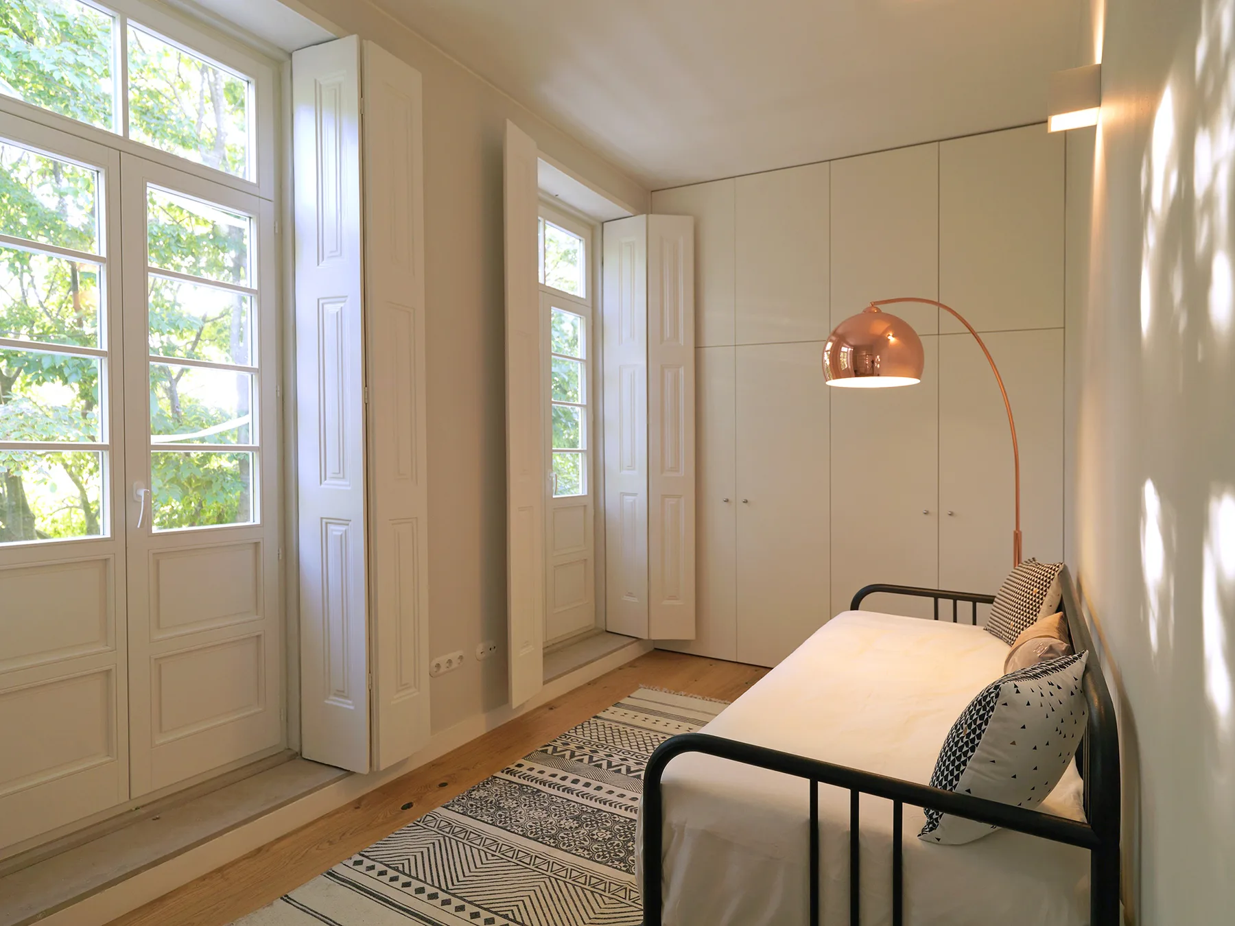 A bright bedroom with a bed, tall windows, and a patterned rug.