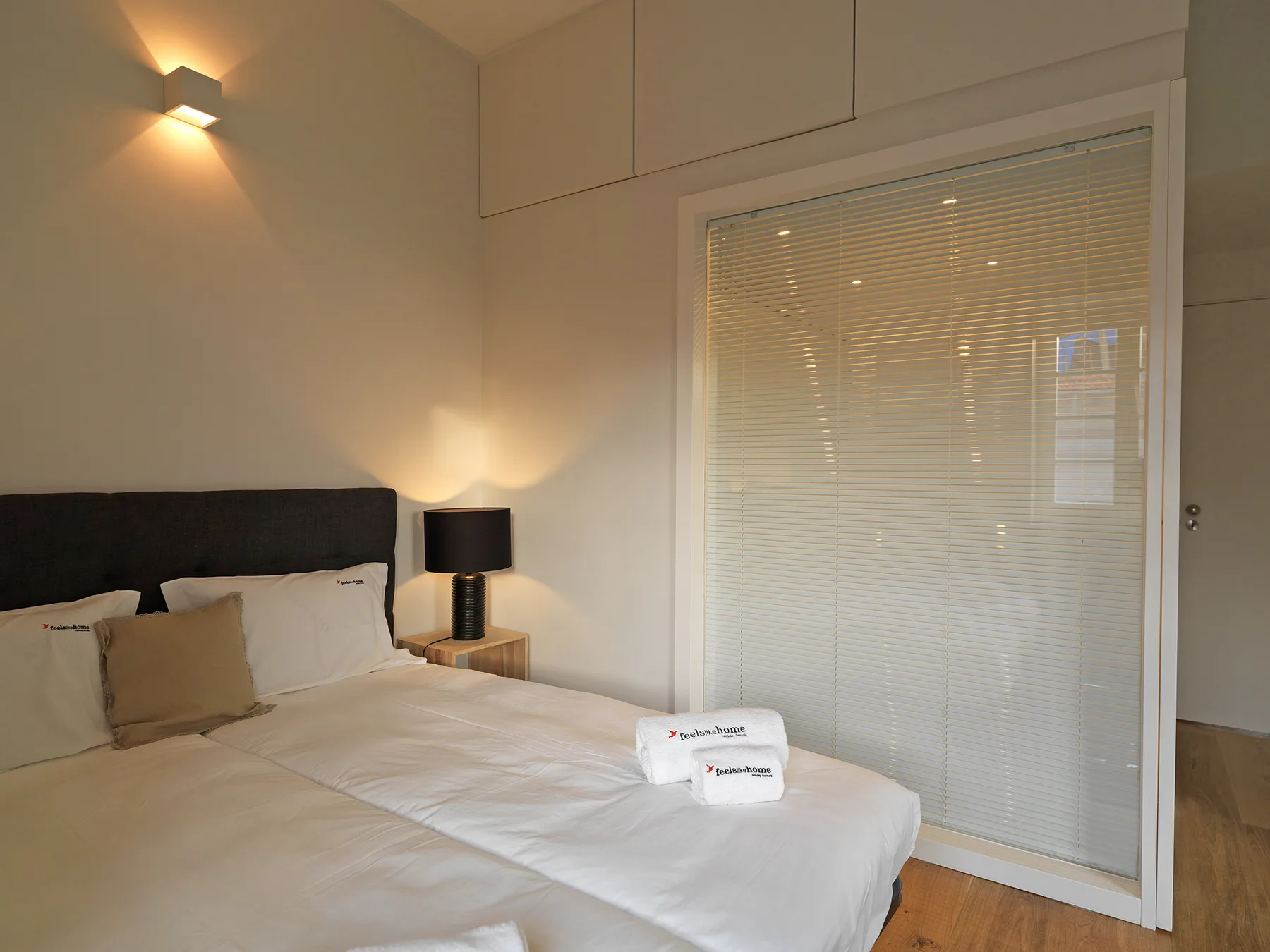 A bedroom with a neatly made bed and a large window with blinds.