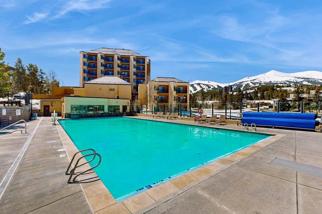 An outdoor swimming pool sits beside a hotel with snowy mountains in the background.