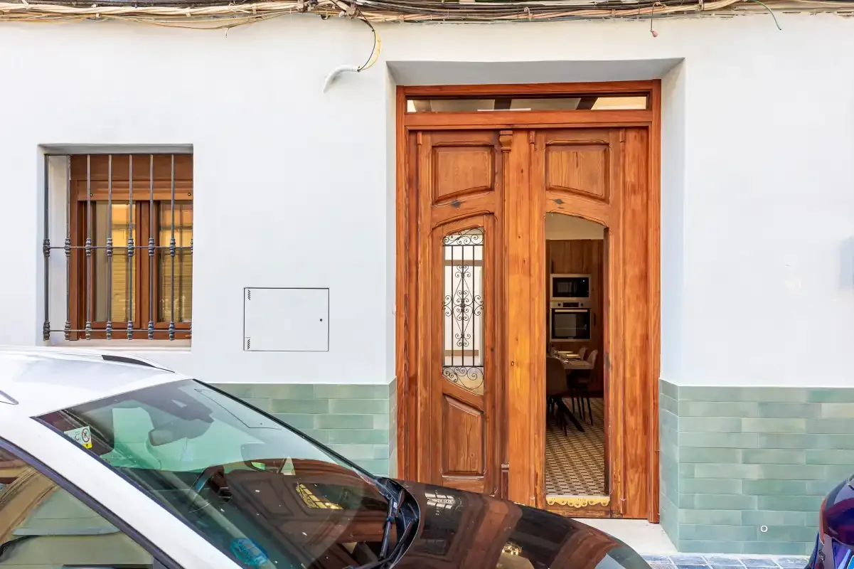 A wooden entrance door and barred window face a street with parked cars.