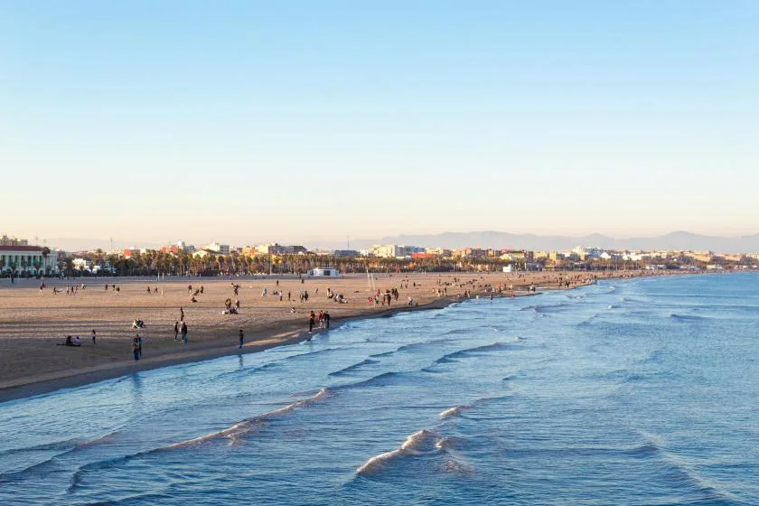 A sandy beach meets calm waves, with people and buildings along the distant shore.