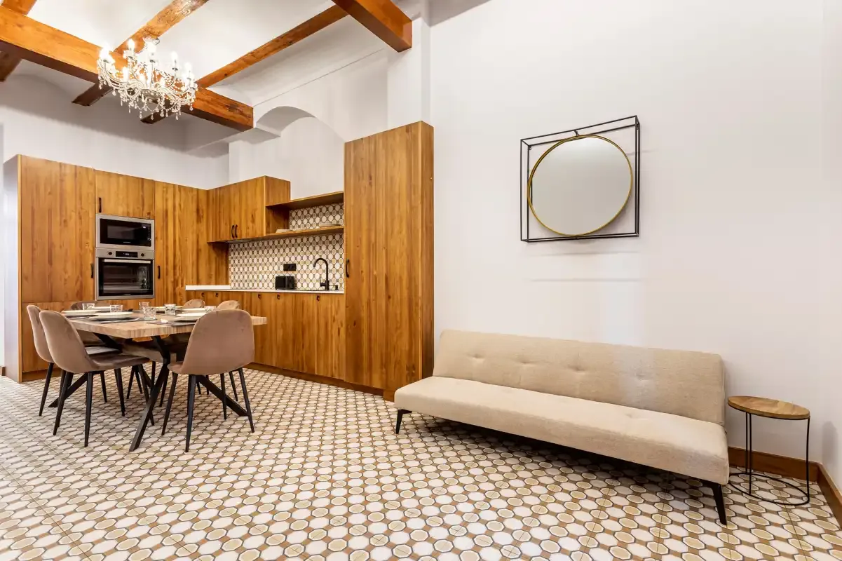 A living and dining area with a beige sofa, wooden kitchen cabinets, and patterned flooring.
