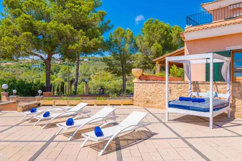 A sunny poolside terrace has lounge seating and a shaded daybed beside a peach-colored building.