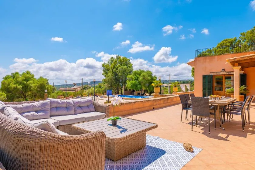 A sunny outdoor terrace has seating, a dining table, and a pool in the background.