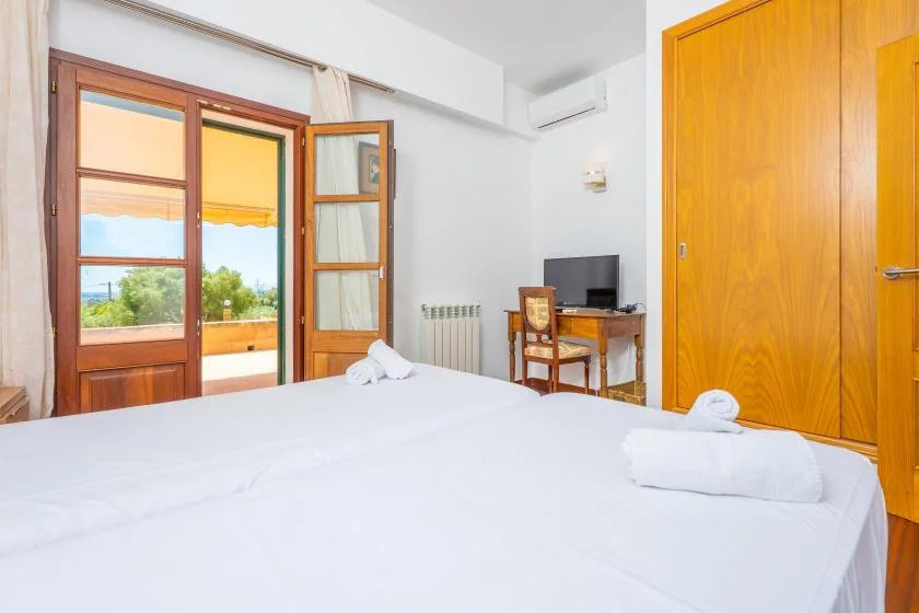 A bedroom with a white bed, wooden doors, and an open window overlooking greenery.