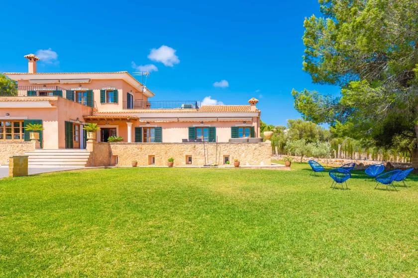 A large house overlooks a green lawn with outdoor seating beneath a bright blue sky.