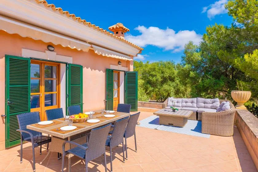 A sunny outdoor terrace with dining seating and a lounge area beside a peach-colored house.