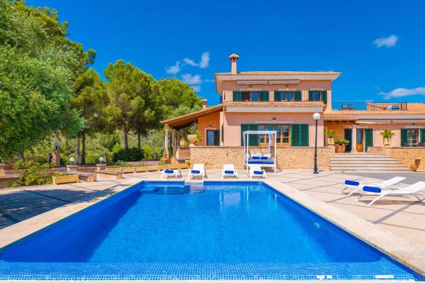 An outdoor swimming pool sits beside a large house and a paved patio.