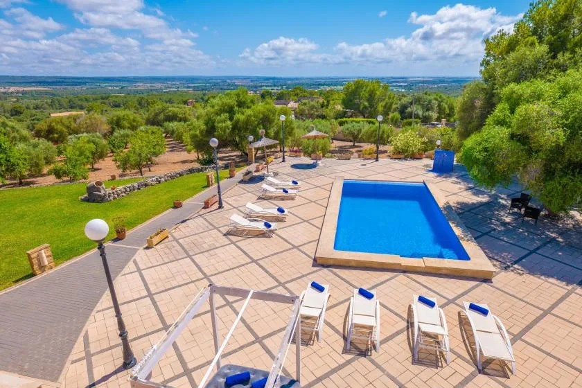 An outdoor swimming pool and sun loungers sit on a tiled patio overlooking green hills.