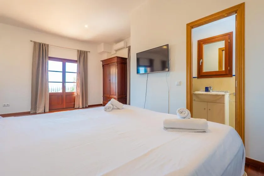A bright bedroom with a large bed, curtained balcony doors, and a wall-mounted television.