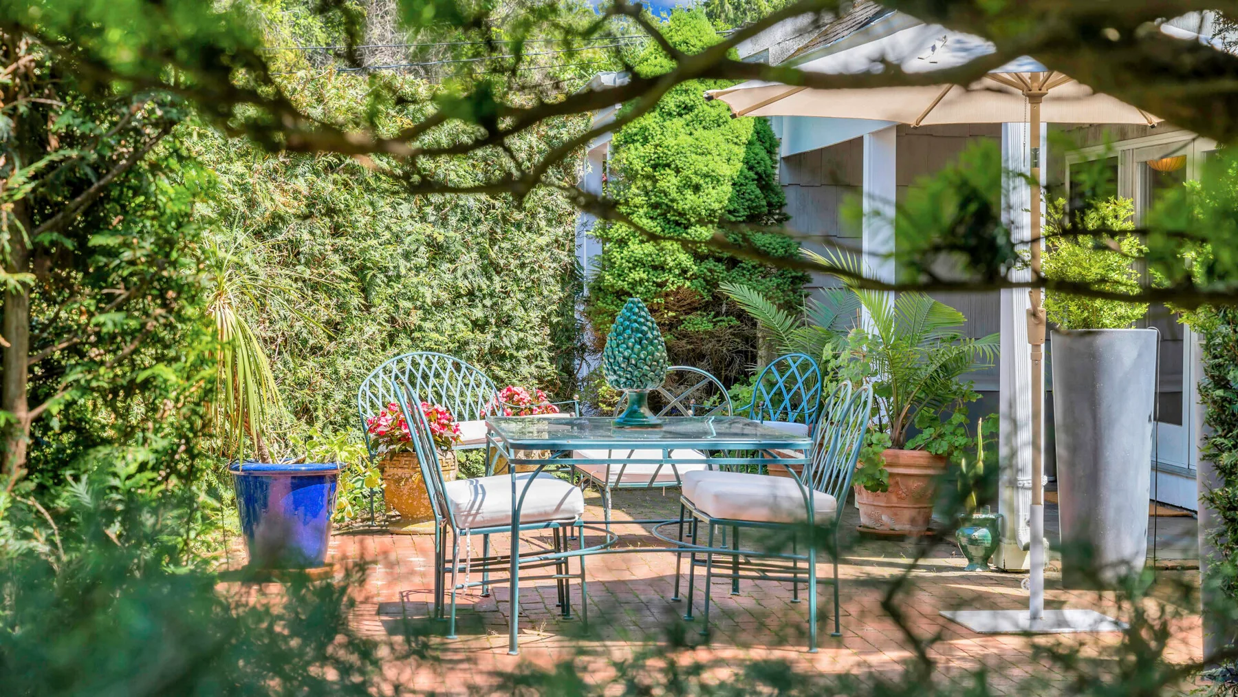 Un patio ajardinado tiene un comedor de metal bajo una sombrilla grande.